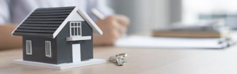 Model house and keys on a table with a person signing documents in the background.