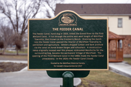 Historical plaque for The Feeder Canal in Wainfleet with buildings and leafless trees in the background.