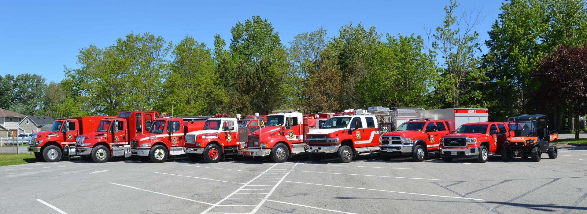 Lineup of red fire trucks and emergency vehicles parked in an open area with trees in the background.