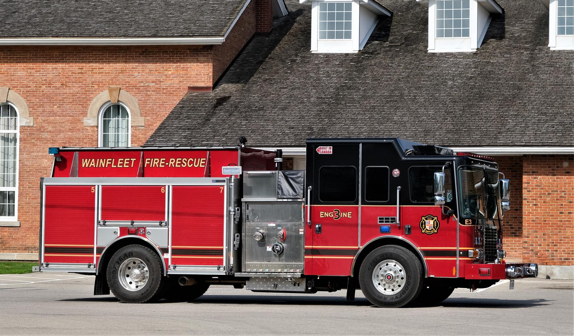 Red and black Wainfleet Fire-Rescue fire truck, labeled "Engine 3," parked in front of a brick building with arched windows. The fire truck is equipped with various tools and compartments.
