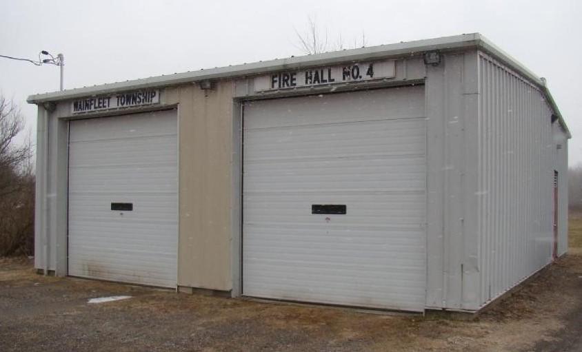 A small, metal-clad fire station building with two garage doors labeled "Wainfleet Township Fire Hall No. 4." The building is grey with a flat roof, set in a rural area with a gravel driveway and bare trees in the background.
