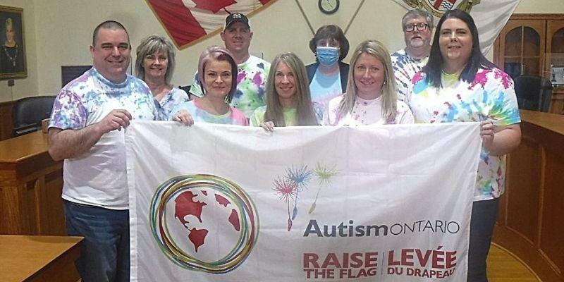 A group of people wearing tie-dye shirts, standing in a room with Canadian flags, holding a banner that reads "Autism Ontario Raise the Flag."
